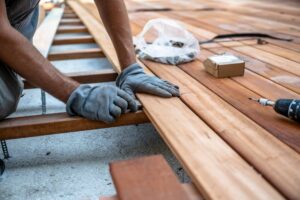 A worker wearing gloves is carefully aligning wooden planks while building a deck, with tools and materials like a drill and screws visible on the surface.